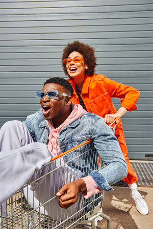 Smiling young african american woman in bright outfit and sunglasses having fun with stylish and scared best friend sitting in shopping cart near building on urban street, friends with stylish vibeの写真素材