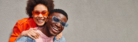 Cheerful african american woman in modern sunglasses and bright outfit hugging best friend and looking at camera while standing near building on urban street, friends with trendy aesthetic, bannerの写真素材