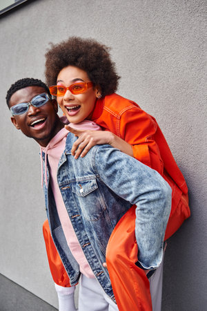 Cheerful young african american woman in stylish sunglasses pointing with finger while piggybacking on best friend and standing near building on urban street, friends with trendy aestheticの写真素材