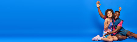 Cheerful young african american best friends in bright summer outfits waving hand and showing yes gesture at camera while sitting on blue background, stylish friends posing confidently, bannerの写真素材