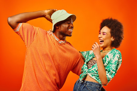Cheerful young african american man in panama hat hugging best friend with bold makeup and summer...