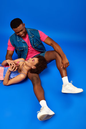 High angle view of positive young african american woman with natural hair and bold makeup lying near best friend in denim vest on blue background, stylish friends posing confidentlyの写真素材