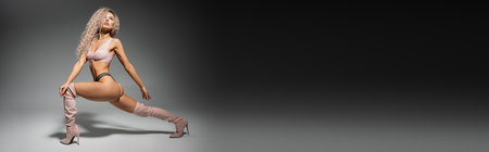 full length of fashion-forward and sexy woman in lace lingerie and long boots standing in expressive pose on black and grey background, toned body, wavy ash blonde hair, banner with copy spaceの写真素材