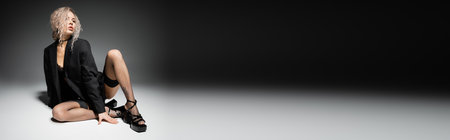 full length of fashionable and seductive woman with ash blonde hair and slender body sitting in blazer, stockings and heeled sandals on black and grey background, sexy style, banner with copy spaceの写真素材