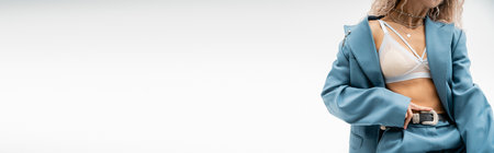 partial view of seductive woman in silver necklaces, bra and blue oversize suit touching leather belt while standing on grey background, sexy look, modern fashion, banner with copy spaceの写真素材