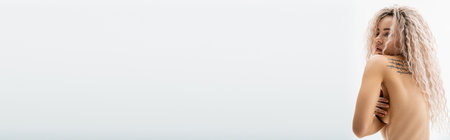 style and sensuality, sexy and charming tattooed woman with shirtless body and wavy ash blonde hair obscuring naked breast with hands and looking away on grey background, banner with copy spaceの写真素材