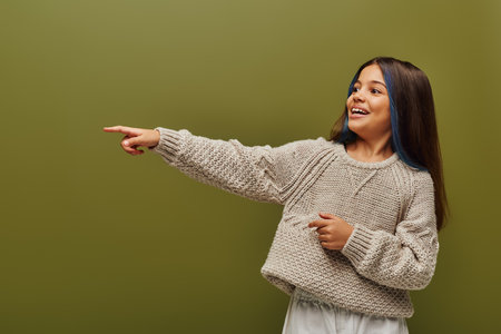 Cheerful and trendy preteen girl with dyed stands of hair wearing modern knitted sweater and pointing with finger isolated on green, contemporary fashion for preteen conceptの写真素材