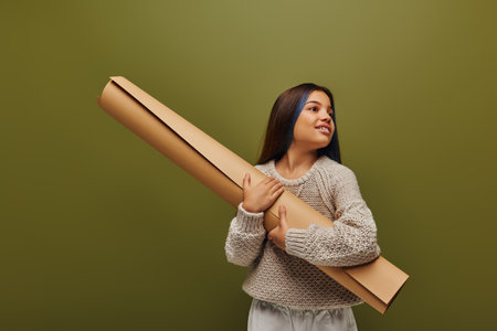 Smiling preadolescent girl with dyed hair wearing autumn outfit and knitted sweater while looking away and holding rolled paper isolated on green, girl radiating autumn vibesの写真素材
