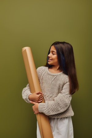Joyful preadolescent with dyed hair wearing stylish and cozy autumn knitted sweater looking at rolled paper and standing isolated on green, girl radiating autumn vibes conceptの写真素材