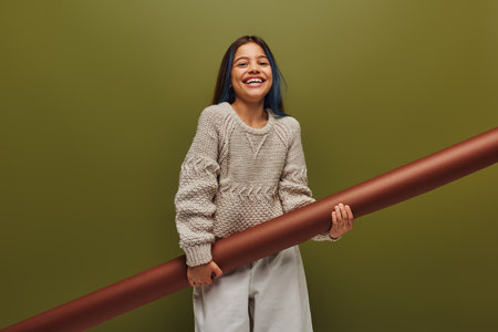 Joyful and smiling preadolescent girl with dyed hair wearing stylish autumn outfit while holding rolled paper and looking at camera isolated on green, girl radiating autumn vibes conceptの写真素材