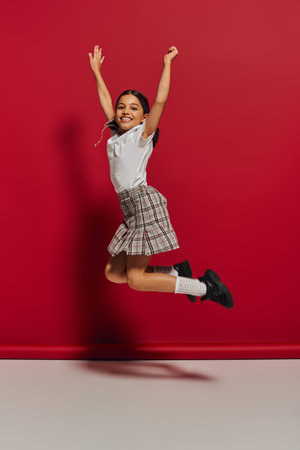 Positive and trendy brunette preteen girl in t-shirt and checkered skirt jumping while having fun and looking at camera on red background, hairstyle and trendy accessories conceptの写真素材