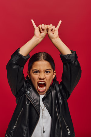 Portrait of mad and fashionable preadolescent girl in leather jacket screaming at camera and showing rock gesture while standing isolated on red, girl with cool and contemporary lookの写真素材