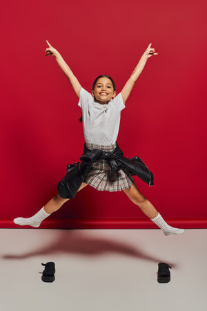 Cheerful and trendy preteen girl in plaid skirt, t-shirt and leather jacket pointing with fingers while jumping and looking at camera on red background, hairstyle and trendy accessories conceptの写真素材