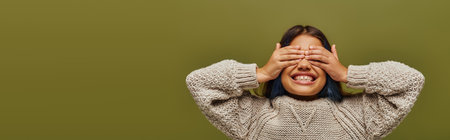 Positive and trendy preteen kid with dyed hair wearing knitted sweater while covering face with...