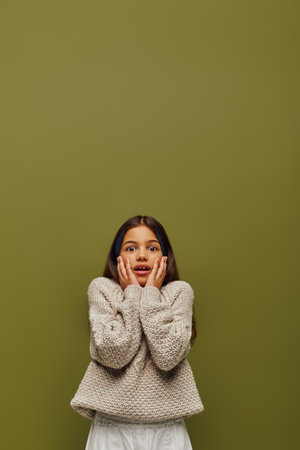 Shocked brunette preteen girl with dyed hair in casual and stylish knitted sweater touching cheeks...