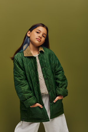 Confident and stylish brunette preteen girl with dyed hair posing in autumn outfit and knitted sweater and looking at camera isolated on green, modern fall fashion for preteens conceptの写真素材