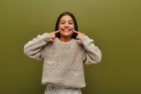 Smiling brunette preteen girl with dyed hair wearing stylish knitted sweater while pointing with...