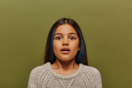 Portrait of scared and stylish preteen girl with dyed hair wearing knitted sweater and looking at camera while standing and posing isolated on green, modern preteen fashionの写真素材