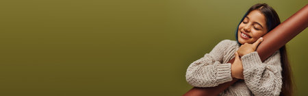 Cheerful and stylish preadolescent girl with dyed hair wearing knitted sweater while hugging rolled paper and standing isolated on green, girl radiating autumn vibes, banner with copy spaceの写真素材