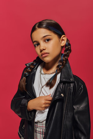 Portrait of confident and trendy preadolescent girl with dyed hair and hairstyle posing in necklaces and leather jacket while standing isolated on red, hairstyle and trendy accessories conceptの写真素材