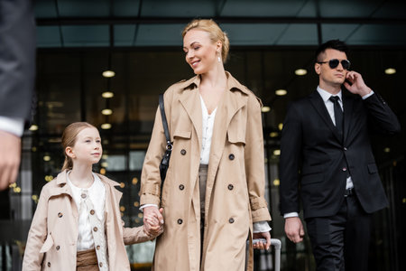 professional bodyguards protecting happy woman and preteen kid, successful blonde mother and daughter in trench coats standing near hotel, safety and protection, private securityの写真素材