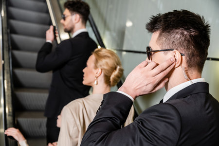 personal security concept, professional bodyguards communicating while protecting safety of clients, rich lifestyle, blonde and successful woman and preteen girl standing on escalator of hotelの写真素材