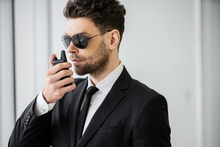 surveillance, handsome bodyguard communicating through walkie talkie, man in sunglasses and black...