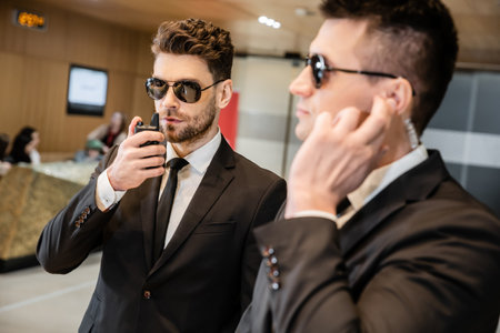 bodyguard service, private security, handsome man in sunglasses and formal wear communicating with his working partner by walkie talkie, hotel security, vigilance, professionalsの写真素材