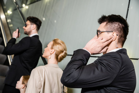 private security, professional bodyguards communicating while protecting safety of clients, rich lifestyle, blonde and successful woman and preteen kid standing on escalator of hotelの写真素材