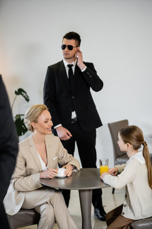 private security concept, successful happy mother and daughter spending time together in cafe, drinking coffee and orange juice, two bodyguards protecting safety of female clientsの写真素材