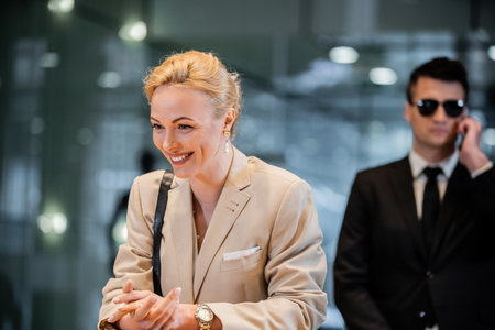 happy blonde woman in formal wear standing at reception desk, personal security service concept, bodyguard in suit on blurred background, hotel industry, luxury travel, formal wearの写真素材