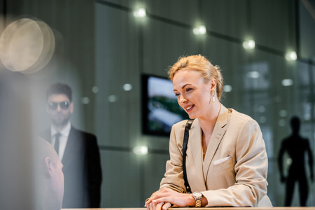 polite businesswoman talking to receptionist in hotel, hospitality industry, blonde and cheerful woman communicating with hotel staff, personal security, private safety, bodyguards on backgroundの写真素材