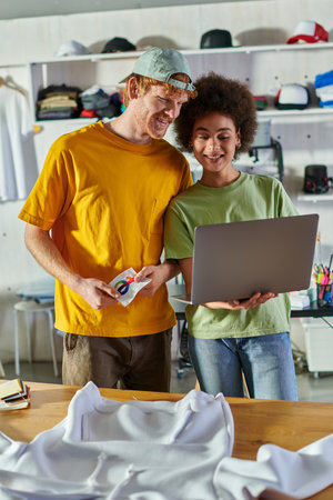 Smiling young african american designer using laptop near colleague with printing layer and clothes on table in blurred print studio at background, self-employment opportunity conceptの写真素材