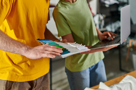 Cropped view of young craftsman holding cloth swatches while working with blurred african american colleague using laptop and standing in print studio, small business conceptの写真素材