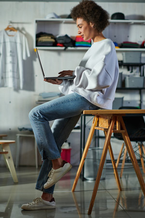 Young african american craftswoman in jeans and sweatshirt using laptop and sitting on table while working in blurred print studio, focused business owner managing workshopの写真素材