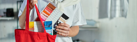 Cropped view of young craftsman in casual clothes putting cloth samples in shoulder bag in blurred print studio at background, self-made success concept, bannerの写真素材