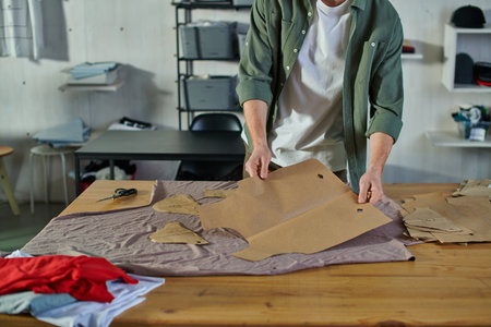 Cropped view of young artisan holding sewing pattern near fabric and scissors on table in blurred...
