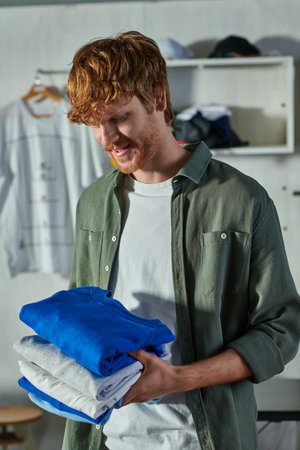 Cheerful young redhead craftsman holding clothes while standing and working in blurred print studio...