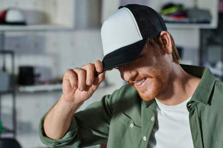 Smiling young redhead craftsman wearing snapback while working and standing in blurred print studio at background, customer-focused entrepreneur conceptの写真素材
