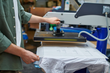Cropped view of craftsman holding layer on t-shirt and working with screen printing machine in blurred workshop, customer-focused small business conceptの写真素材