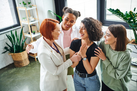 redhead motivation coach and smiling multicultural female friends embracing overjoyed multiracial woman during psychology session in consulting room, moral support and mental wellness conceptの写真素材