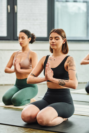 young tattooed woman sitting in thunderbolt pose and meditating with closed eyes and praying hands...