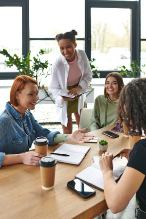 smiling tattooed woman pointing with hand while talking to multicultural team of interest club near table with smartphones and takeaway drinks, mutual support and understanding conceptの写真素材