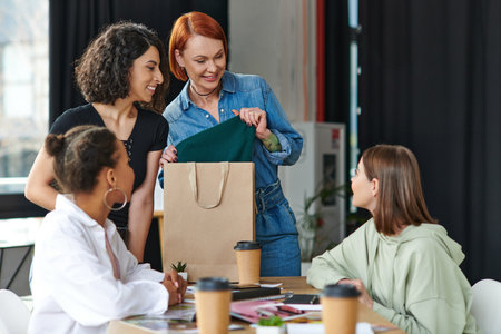 redhead woman with shopping bag showing purchase to smiling multiethnic girlfriends near paper cups...