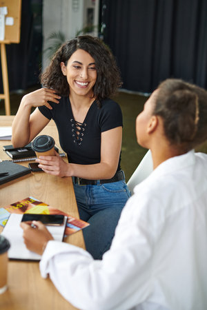 joyful multiethnic woman talking to african american friend while sitting with coffee to go near...