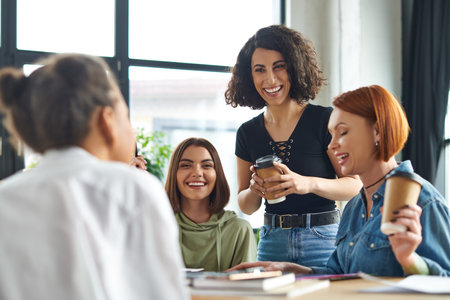 excited multiracial woman standing with paper cup of coffee to go during conversation with multiethnic female friends in interest club, spending time in friendly diverse communityの写真素材
