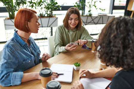young and smiling woman networking on mobile phone, spending time with friends sitting at table with...