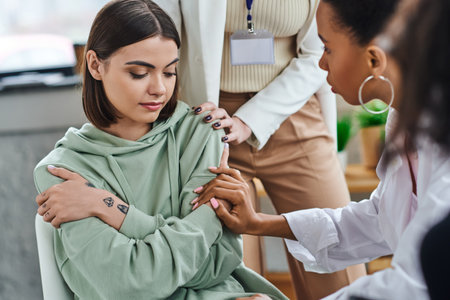 professional psychologist and african american girlfriend touching depressed and offended tattooed woman sitting with folded arms during supportive therapy, empathy and solidarity conceptの写真素材