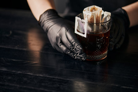 pour-over brewing method, glass with coffee in paper filter bag near barista in black latex gloves