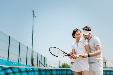 happy man teaching girlfriend how to play tennis on court, holding rackets and ball, sport and funの写真素材